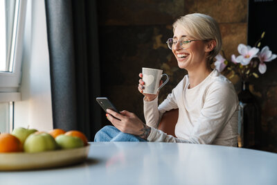 Woman laughing while drinking tea and reading her phone screen