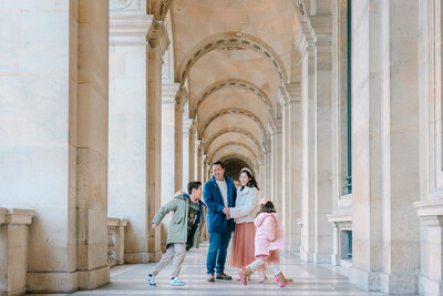 Playful family of four — mother, father, son, and daughter — running around and laughing near the Louvre Pyramid and Palais Royal during a Paris family photoshoot.
