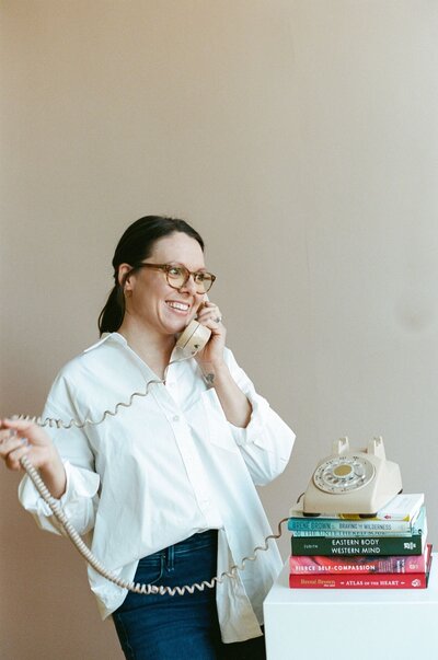 A woman named Kristen Simons, wearing glasses, talks on the phone surrounded by a stack of books.