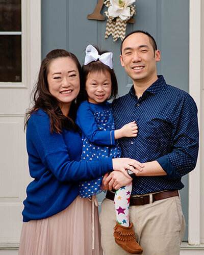 A family standing in front of their front door. Dad is holding a little girl and mom has her arms wrapped around them.
