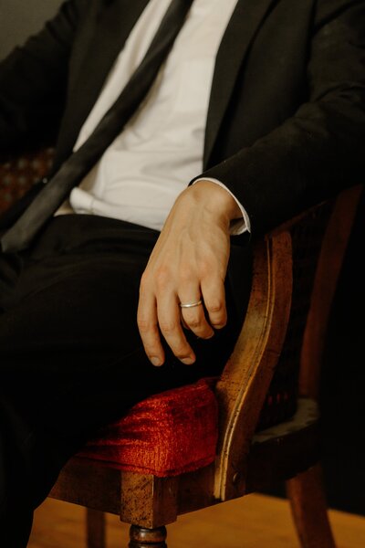 A groom lounges in a vintage velvet chair showing off his new wedding ring.