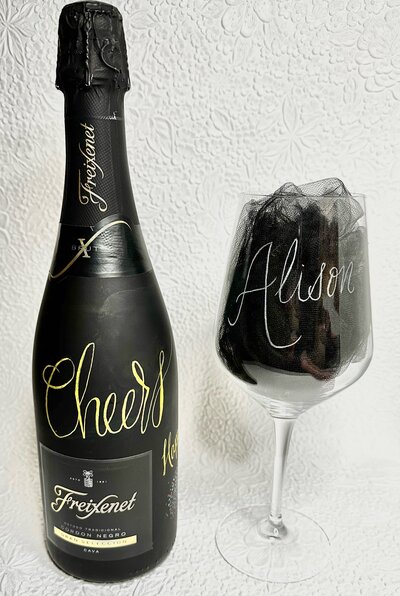 A matte black bottle of freixenet prosecco that has "Cheers" written in metalic gold calligraphy beside a stemmed wine glass that has been hand engraved to say "Alison".