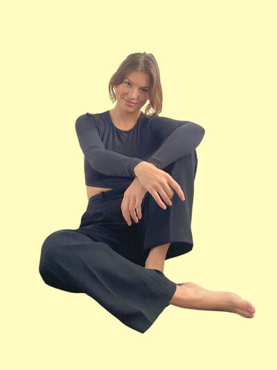 Caitlin Christensen sits cross-legged on the floor with a relaxed smile. She wears a black long-sleeve crop top and black trousers against a soft yellow background.