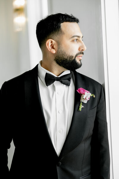 Groom in classic black tuxedo with pink boutonniere looking out window at Maison De Veaux wedding venue in Houston, Texas during morning preparations