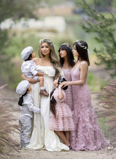 A mother in an ivory off the shoulder dress surrounded by her five children with her girls all wearing dusty rose dresses and her boys in grey linen outfits with all the females wearing baby's breath floral crowns on their heads and posing in a manner that is similar to classical paintings of people.