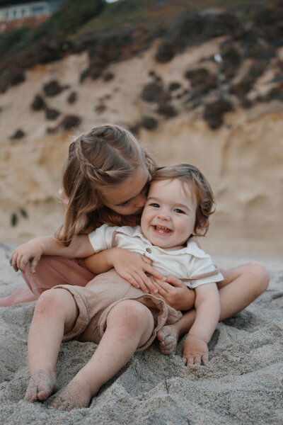 family of four sitting and standing on sand dunes, smiling at each other during a sunny beach day