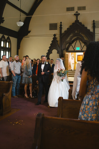 Bride walking down the aisle with her father during a church wedding ceremony, holding a bouquet and surrounded by guests.