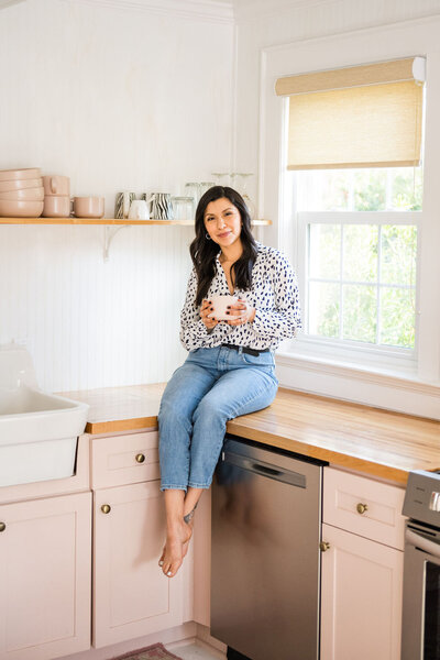 real estate agent, karen wearing a black and white blouse and light jeans sitting on the counter in the kitchen next to the sink  holding a white coffee cup  half smiling