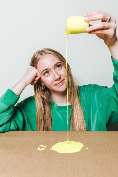 Woman with long blonde hair wearing a green long-sleeved shirt pours bright yellow paint on brown craft paper.