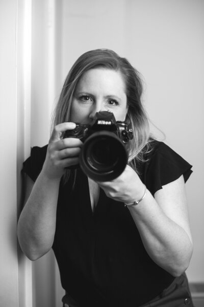 Black and white headshot photo of Boston personal branding photographer Kristin Palkoner, owner of Lens and Prism Photography holding her Nikon camera.