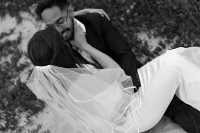 Black and white photo of a bride in a veil sitting on her groom’s lap as they smile at each other outdoors.