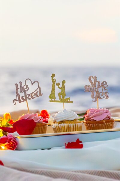 Close-up of proposal cupcakes on a sailboat in Hawaii decorated with He Asked and She Said Yes toppers, surrounded by rose petals and ocean views.