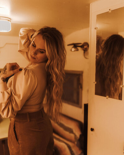Dani Canerday hair extension specialist in dallas stands in front of a full-length mirror dressed in a beige blouse and brown pants. She is in profile showing her wavy, lightning blonde, long, wavy hair.
