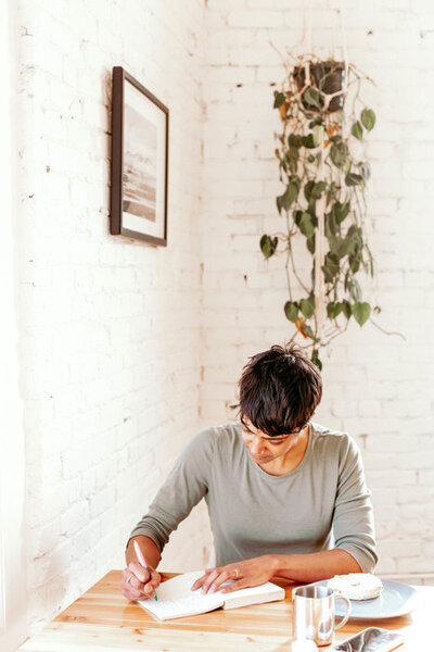 Woman with black pixie cut takes notes in her notebook in front of white brick wall and hanging plant
