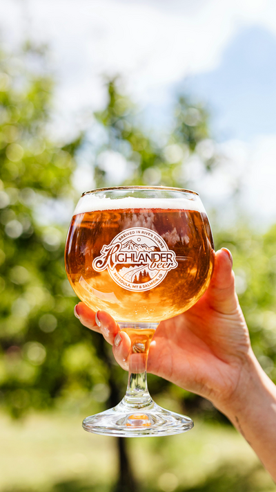 A hand toasts a glass of draft beer that has a decal of the Highlander Beer logo illustration, a mountain scene with a sun, forest, and river running through it as well as the text "Highlander Beer"