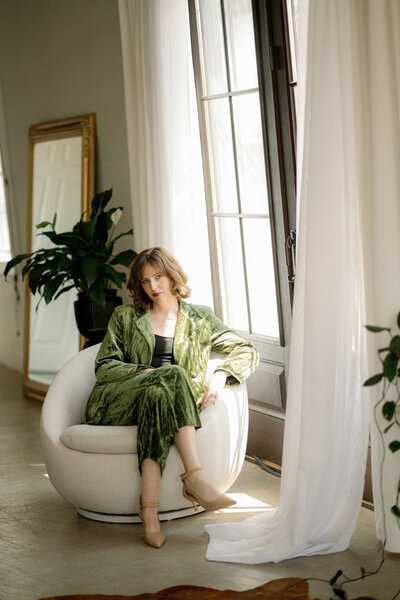 A woman in a green velvet suit sits in a white upholstered chair next to tall wooden doors with a houseplant and ornate gold-framed mirror in the background