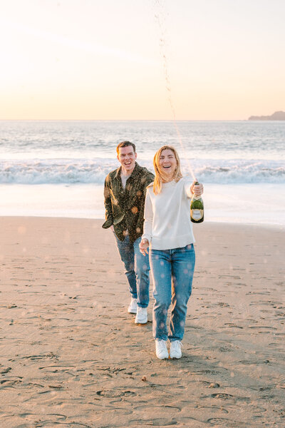 engaged couple laughing and popping a bottle of champagne on the beach dressed in casual jeans and sweaters.
