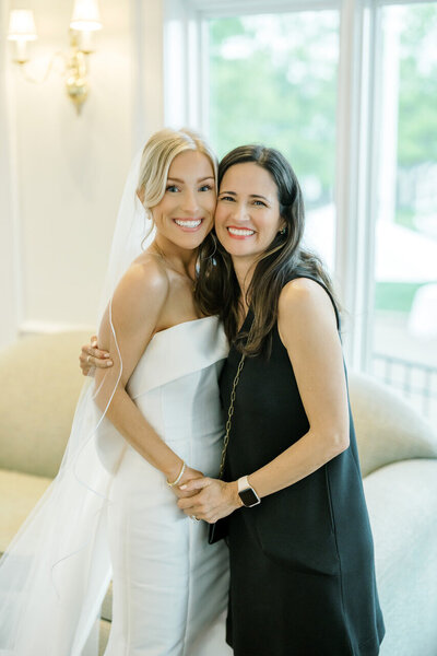 Bride in a white gown and veil smiling beside another woman in a black dress