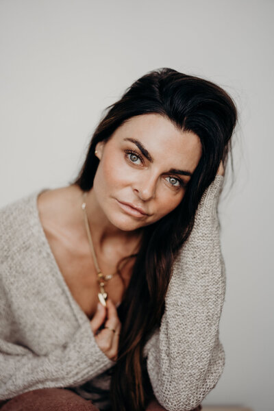 A striking dark haired, blue eyed woman makes direct eye contact while softly smiling as she holds on to a unique gold necklace while wearing and oatmeal colored sweater.