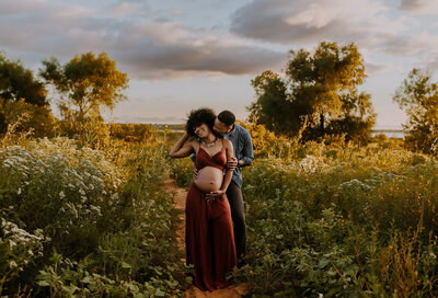 A bare belly pregnant woman in a two-piece set is standing on a trail at Murrell Park in Flower Mound, Texas with her husband as he leans over her shoulder and kisses her neck and she laughs