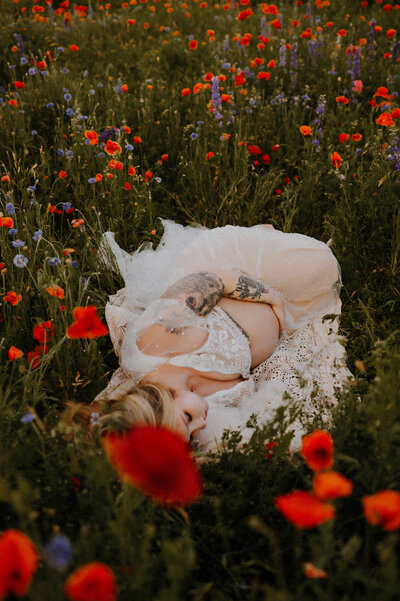 An expecting mother in a lacy white set laying in a field of wildflowers.