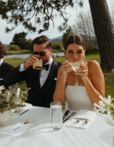 The bride and groom sit side by side under the open sky, sharing a quiet toast with their signature cocktails—soaking in the joy of the day, one sip at a time.
