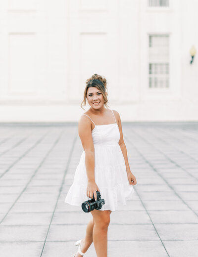 Abby, the owner of Abby Bell Photography, fine art wedding and portrait photographer in Alabama, smiling while holding her camera in an elegant white dress.