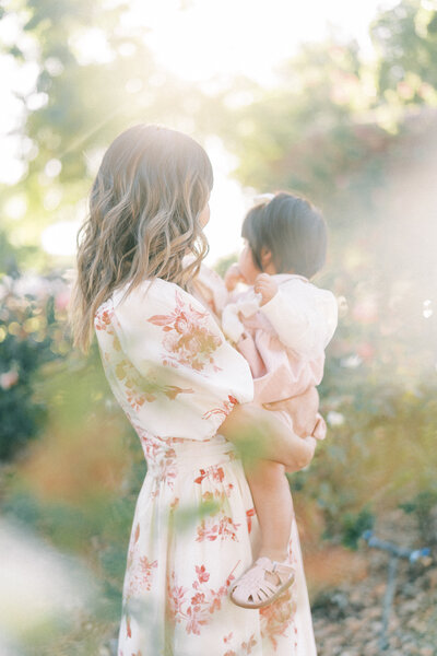 mom in a white dress dress with pink flowers on it holding her one year old daughter in a rose garden with the sun shining behind them.