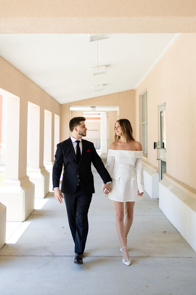 A bride and groom walk hand in hand through the breezeway of the St. Johns County Courthouse in St. Augustine after their courthouse wedding. The bride wears a chic off-the-shoulder dress, and the groom is dressed in a classic black suit. Captured by a St. Augustine elopement photographer, this moment reflects the elegance and simplicity of a courthouse elopement at the St. Johns County Courthouse Wedding Chapel.