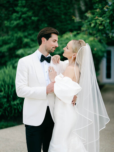 Bride and Groom looking at each other. Groom is wearing a white tux jacket with a black bowtie  and black trousers and the bride is wearing a dress with puff sleeves and her veil is satin trimmed.