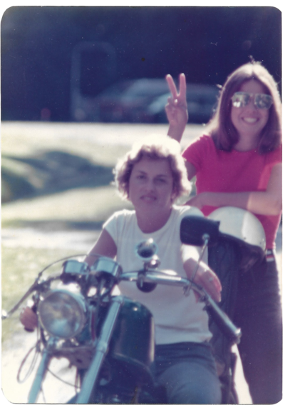 one sister sitting on a motorcycle and the other has sunglasses on and is holding up a peace sign with her hands, in the 1970s. 