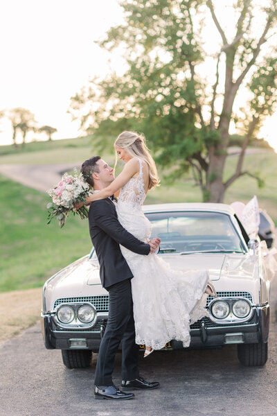Bride and groom celebrating with a lift in front of a vintage classic car at sunset, photographed by Zoe Evans Photography.