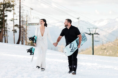 A bride and groom walk up a snowy slope while carrying their snowboards in Mammoth Lakes