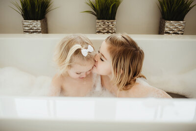 Big sister kissing little sister on the temple while in the bubble bath.