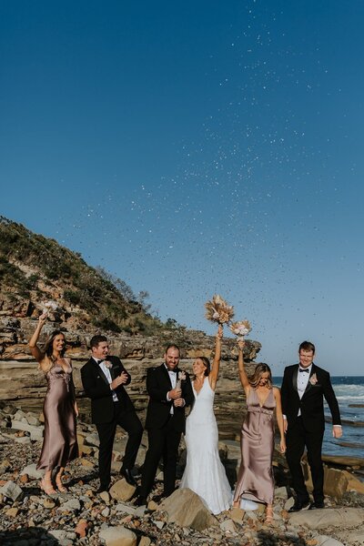 Colourful image of a wedding party firing a confetti canon in the air whilst standing on the rocks at Terrigal Beach with the ocean behind. Central Coast Wedding Photographer