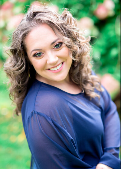 headshot portrait of woman standing outside wearing a blue long-sleeved blouse & smiling, posing for a South Shore, MA portrait photographer
