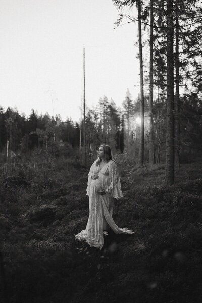 Svartvit bild på en gravid kvinna i vit klänning som står i skogen.