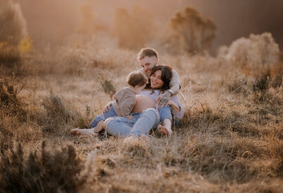 Family sitting in a sun-drenched field at golden hour, toddler is kissing his mothers pregnant belly.