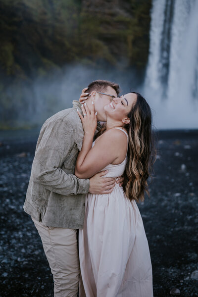 Joyful couple shares a kiss near Skógafoss waterfall, wrapped in love and mist in Iceland’s dramatic landscape.