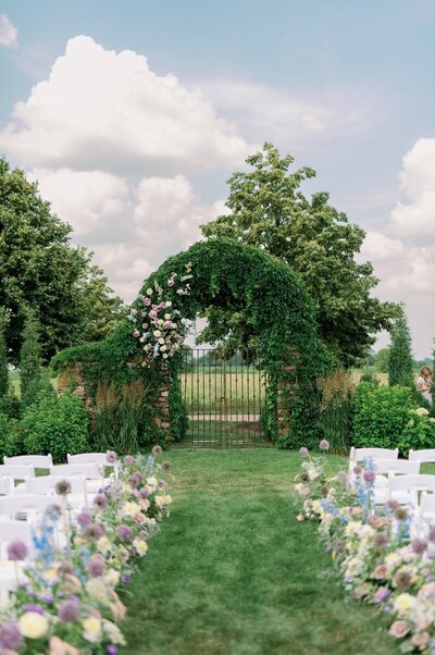 outdoor wedding set up in minneapolis minnesota with gate surrounded by lush green archway and white chairs adorned with flowers