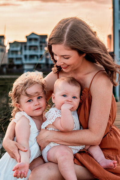 A woman is holding a young baby and has her arm around an older toddler. The two girls are looking at the camera with cute looks on their faces while the woman looks down at them smiling.