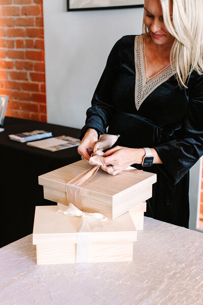 A woman in a black dress ties a ribbon on a gift box at SoHo63, a Chandler wedding and event venue. She stands near a brick wall and a table with stacked boxes, highlighting the venue’s elegant and stylish indoor spaces perfect for events and weddings.