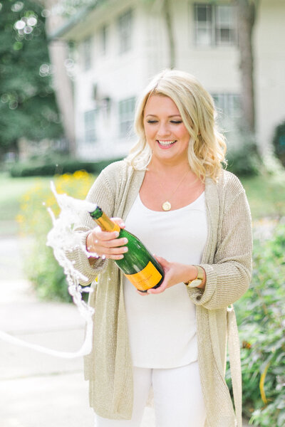 woman smiling and popping open champagne bottle, wearing a gold cardigan and a white t-shirt and a gold necklace