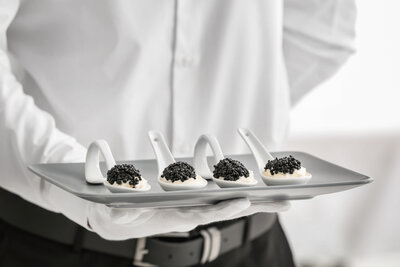 White spoons of caviar being served on a tray held by a server wearing a white shirt, white gloves, and black belt