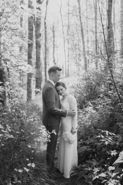 Bride and groom are standing in a forest and bride is leaning her head against his chest in Finland