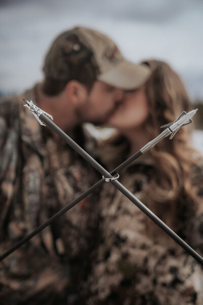 A couple share a kiss in the background while holding two archery arrows crisscrossed in the foreground, an engagement ring hooked on them both.