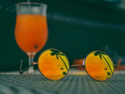 A pair of round, yellow-tinted sunglasses resting on a textured surface, reflecting the silhouette of palm trees and a vibrant sunset. In the background, a tropical orange cocktail in a glass adds to the ambiance. This inviting image represents the relaxing and rewarding experiences that vacation fundraising for charities can provide to supporters while advancing impactful causes.
