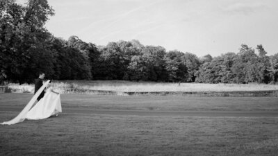 A bride and groom walk across a lawn in a documentary-style black and white photo.