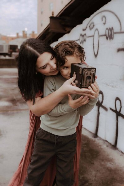 A woman helps a young boy hold and look through a vintage camera on a rooftop with graffiti behind them during a Warner Robins Urban Family session by Fire Family Photography.
