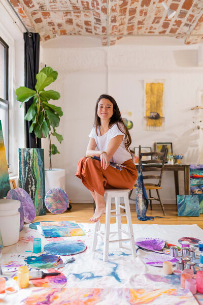 Courtney sitting on a stool in her creative studio workspace with artist surrounded by colorful paintings and supplies
